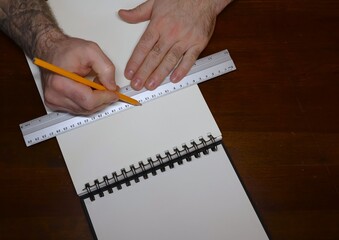 A large notebook, two male hands holding a pencil and a metal ruler are visible on a brown table.