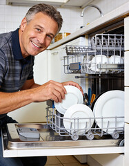 Mature Man Washing Dishes In Dishwasher In Home Kitchen
