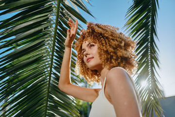 curly woman with natural hair enjoys sunny day outdoors under tropical palm leaves in white...
