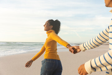 Cheerful young woman walking while holding hand of boyfriend at beach during sunset