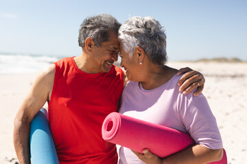 Happy senior couple holding yoga mats touching foreheads of each other at beach