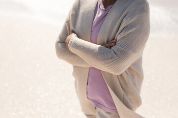 Midsection of senior woman with arms crossed standing at beach on sunny day