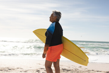 Side view of senior woman carrying surfboard looking away at beach during sunny day