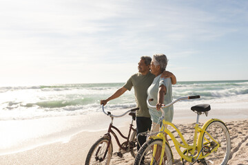 Happy multiracial senior couple looking away standing with bikes at sunny beach against sky © wavebreak3