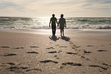 Senior multiracial couple hold hands, walking on sunny beach with footprints