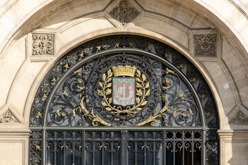 Obraz premium Intricate wrought ironwork with gold leaf motifs and a detailed civic crest adorn the grand arched entrance of Hotel de Ville in Paris, highlighted by ornate stone carvings and bright natural light.
