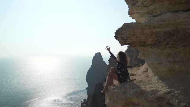 Woman cliff sea, young female tourist sits dangerously on a sheer rock edge overlooking the vast sparkling blue ocean under bright sunlight.