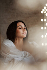 A woman with black hair wakes up in the morning in a white blanket and catches the rays of the sun. The portrait is creative, with brown eyes. High quality photo