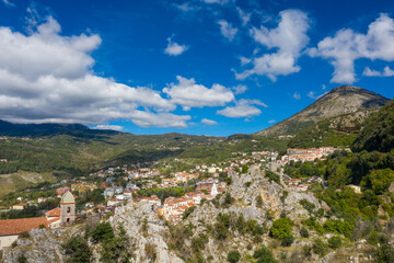 Obraz premium Picturesque view of Lauria, Italy, nestled among rugged rocky outcrops and lush green hills under a vibrant blue sky with scattered clouds. Sunlight highlights the colorful houses and dramatic