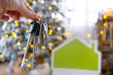 Keys in hand on background fairy lights Christmas tree and tiny house gift and cat for New Year, Christmas. Building, design, project, moving, insurance, mortgage, rent and purchase real estate.