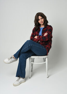 Full length portrait pretty young brunette female model wearing casual modern plaid  flannel shirt and jeans, sneakers. Sitting in chair, relaxed Isolated figure posing on white studio background.