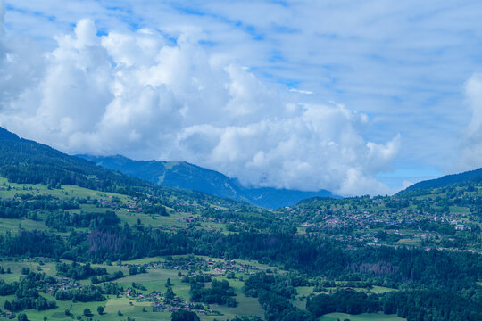 Expansive view of rolling green hills dotted with scattered alpine homes and villages in Combloux, framed by dramatic cumulus clouds and distant blue mountains.