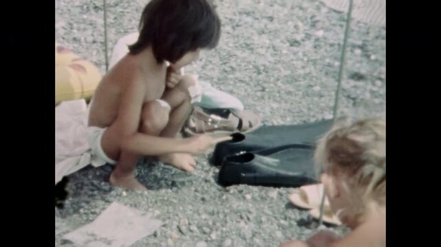 Mischievous child playing funny prank by putting crab into father beach sandal, swimming fins. Playful childhood humor in sea vacation. Summer fun, jokes on holiday. Old film. Archival retro archive