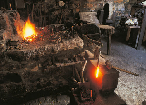 Interior of a traditional blacksmith workshop featuring an active forge and a glowing hot iron piece on an anvil. The scene highlights craftsmanship, heat, and manual metalworking in a rustic setting. - Powered by Adobe