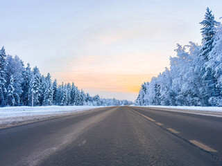 Beautiful winter road through a snowy forest at sunset, scenic asphalt highway with frost covered trees, winter landscape with glowing sky and hoarfrost