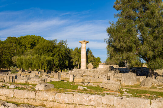 Ancient stone remains and a standing column from the Temple of Zeus rise among lush trees and scattered ruins at Olympia, Greece. Warm afternoon light highlights the textured masonry and vibrant