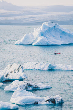 View of a lone kayaker navigating through the serene, icy waters amidst colossal icebergs, their textures a stark contrast against the water, Hofn, Vatnajokull, Iceland.