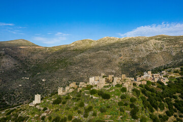 Obraz premium Aerial view of the historic stone village of Vathia perched on a lush hillside, surrounded by rugged mountains and clear blue sky. The old towers and clustered buildings create a striking contrast