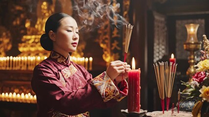 A traditional Chinese woman lights incense sticks from a candle in a temple. Praying during a spiritual ceremony for Lunar New Year. Asian cultural and religious ritual