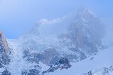Fototapeta premium A veil of mist softens the dramatic snowfields and rugged rock faces of Mont Maudit, with icy blue glacier textures emerging through the haze in the alpine landscape.