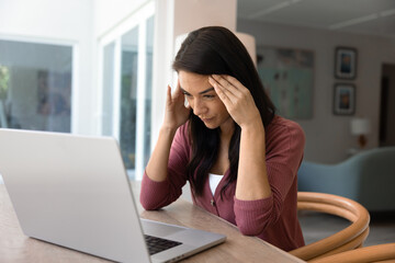 Woman sitting at table with laptop, holding her head in frustration due to device malware,...