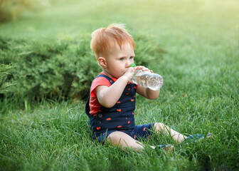 Little boy is sitting on the grass and drinking water in the park in the summer. Child is thirsty. Protection kids from insects