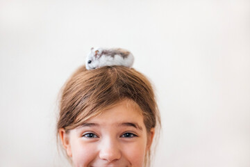 Young girl with brown hair smiling joyfully, a small hamster perched on her head, showcasing a playful and affectionate bond between child and pet in a bright atmosphere