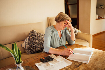 Mature woman with short hair is sitting at a wooden table, calculating expenses with a calculator, surrounded by papers and a plant, reflecting on financial planning and budgeting