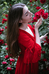 Young woman in a flowing red dress gently touches vibrant red roses in a lush garden, surrounded by greenery, capturing the essence of nature and beauty in harmony
