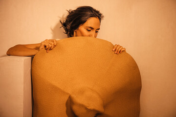 Woman with long hair, holding a large straw hat, poses against a textured wall, showcasing a relaxed and artistic vibe in a warm, inviting atmosphere