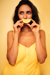 Woman with dark hair wearing a yellow dress playfully holds a slice of lemon to her mouth against a vibrant yellow background, embodying freshness and joy in a cheerful setting