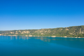 Obraz premium Serene view of Lac de Sainte-Croix with calm blue water reflecting a sunlit forested hillside under a vibrant, cloudless sky.