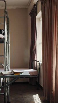 Sunlight streaming through window illuminating rows of identical metal bunk beds and single beds in a room with pink curtains and tiled floor, creating a repetitive and patterned composition