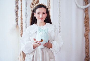 Young woman with long dark hair, wearing a white blouse and pearl accessories, holds a decorative vase in a softly lit room with textured wall hangings, showcasing elegance and charm