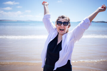 Joyful woman with sunglasses celebrates on sandy beach, arms raised in excitement, with ocean waves and clear blue sky creating a vibrant and uplifting atmosphere of happiness