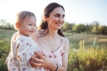 Woman holding a smiling baby girl in a sunlit field, surrounded by greenery, showcasing a joyful moment of connection and love in a natural outdoor setting