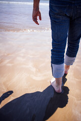 Man walking barefoot along sandy beach shoreline, with waves gently lapping at feet, casting a long shadow on the wet sand, evoking a sense of tranquility and connection to nature