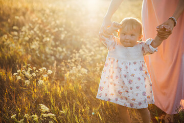 Little girl in floral dress is being held by adult in pink dress, walking through sunlit field with wildflowers, capturing a moment of joy and connection in nature