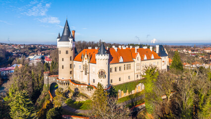 A drone captures Zleby Castle from an aerial perspective. The scene includes surrounding trees and buildings in sunny weather. This location showcases the castle's unique architecture.