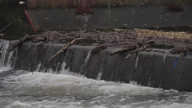 A static close-up shot of white water cascading over the concrete edge of a river weir on the River Wear in Durham, showing accumulated driftwood