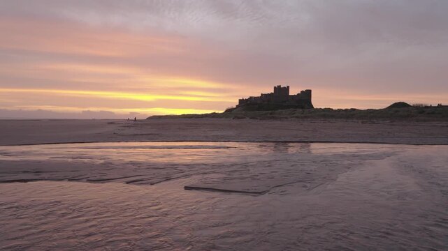 Low, wide clip of Bamburgh Castle in Northumberland. Taken at sunrise from beach with small stream in foreground 