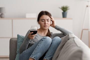 Obraz premium Sad indifferent caucasian young lady sits on sofa with glass of wine and suffering from headache in home interior. Alcohol addiction, alcoholism, hangover, loneliness and depression, mental health