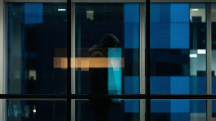 A figure in a hooded jacket stands gazing out of a large window. The city lights outside are reflected in the glass, creating a mesmerizing display of colours and shapes.