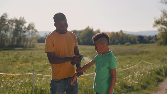 African American man spraying a child with insect repellent to protect his son from mosquito and tick bites in a countryside landscape