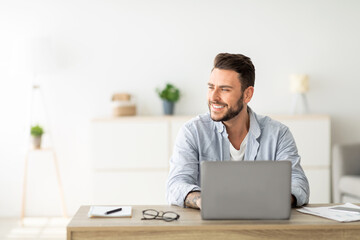 Fototapeta premium Happy freelance man working on laptop in home office, sitting on workplace and looking aside at free space. Smiling guy thinking about successful business strategy or dreaming about holiday