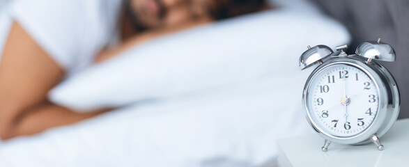 An alarm clock is placed on a nightstand beside a sleeping person in bed. The room is dimly lit,...