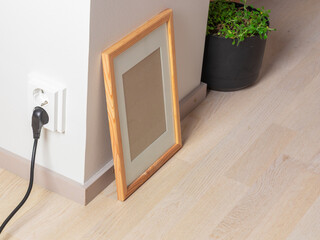 A light wood photo frame leans against a wall next to an electrical outlet and a potted plant,...