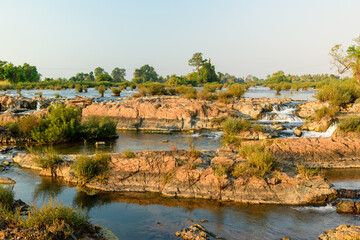 Fototapeta premium Soft morning sunlight highlights layered stone, gentle rapids, and scattered green vegetation at Tat Somphamit Waterfalls, also known as Li Phi Falls, in Champasak Province, southern Laos. The dry