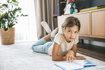A young girl lying on a carpet in a modern living room, focused on reading a book. Childhood education, leisure, and home-based learning in a comfortable domestic environment