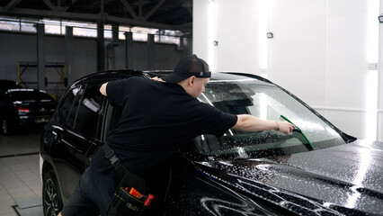 Careful worker applying a paint protection film to the surface of a shiny black car. Automotive detailing service in a clean garage